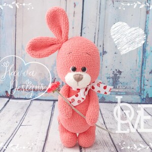 May include: A handmade coral-colored crochet bunny plushie with long ears, holding a red flower. The bunny has a white and red polka-dot bow. The image includes the text "Havva Designs" and the word "LOVE".
