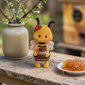 Op de afbeelding: Gehaakte bijenpop met een rode bloem, met een honingpot. De bij heeft zwart-gele strepen en witte vleugels. Een vaas met knoppende takken en een bord met honingraat staan op een houten oppervlak.