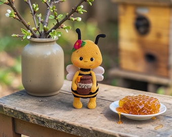Peut inclure: Poupée abeille au crochet jaune et noir, tenant un pot de miel. Un vase avec des branches en bourgeon et une assiette de rayon de miel sont sur une surface en bois. La poupée a une fleur rouge sur la tête.