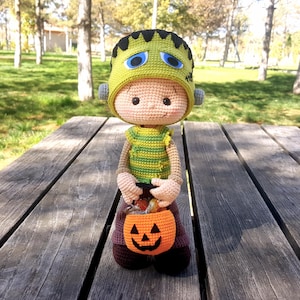 May include: A handmade crochet doll dressed as Frankenstein for Halloween. The doll wears a green and black hat, a green and yellow striped shirt, and brown pants. It holds an orange pumpkin-shaped candy bucket. The doll is sitting on a wooden picnic table.