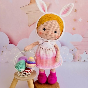 Puede incluir: Muñeca de ganchillo hecha a mano con un gorro de orejas de conejo blanco y un vestido rosa, sosteniendo una cesta con coloridos huevos de Pascua de ganchillo. La muñeca está sobre una base de corcho, con un fondo rosa claro.