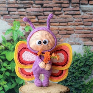 Könnte beinhalten: Handgefertigte Häkelpuppe in Form eines Schmetterlings mit lila Körper, Kopfbedeckung und Antennen. Die Puppe hat große Kulleraugen und hält orangefarbene Blumen. Die Flügel sind gelb, orange und rosa. Die Puppe sitzt auf einer Holzoberfläche.