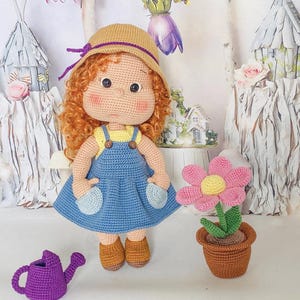 Puede incluir: Muñeca de ganchillo hecha a mano con cabello rizado rojo, vestida con un vestido azul y un sombrero de paja con una banda morada. También hay una regadera morada y una flor rosa en una maceta marrón. Un artículo artesanal.