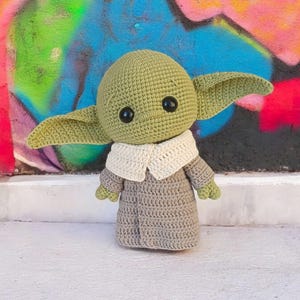 Pode incluir: Boneca de crochê de um personagem com orelhas grandes, pele verde e olhos pretos. A boneca usa uma túnica cinza com gola branca. O fundo é uma parede de grafite colorida.