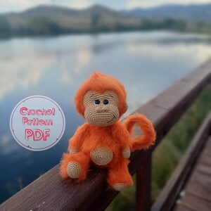 Könnte beinhalten: Eine orangefarbene Häkelpuppe in Form eines Affen mit flauschigem Körper und gehäkeltem Gesicht, Händen und Füßen. Die Puppe hat einen runden, beigefarbenen Bauch und sitzt auf einem Holzgeländer. Der Text "Crochet Pattern PDF" ist enthalten.