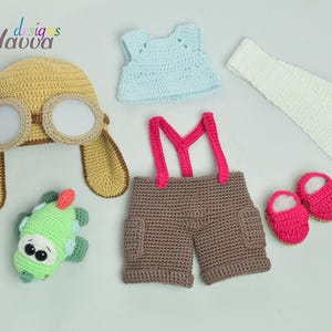 May include: A collection of handmade crochet baby clothing and accessories. Includes a yellow aviator hat with goggles, a light blue top, brown shorts with pink suspenders, white leg warmers, a green toy, and pink booties.