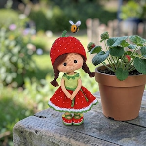 May include: A handmade crochet doll dressed as a strawberry, with a red hat and dress, green top, and brown pigtails. A small bee is perched above the doll. A potted strawberry plant sits nearby.