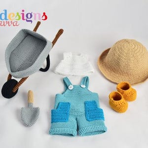 May include: A collection of crocheted baby items, including a gray wheelbarrow, a shovel, a white top, blue overalls with blue pockets, a straw hat, and yellow booties. The items are arranged on a white surface.