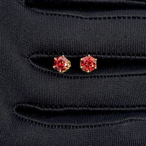 May include: A pair of stud earrings featuring vibrant red gemstones set in gold-colored metal. The gemstones have a faceted, hexagonal shape. The earrings are displayed against a black background.