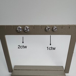May include: A display of two pairs of stud earrings. One pair is labeled "2ctw" and the other "1ctw". The earrings are round with a silver bezel setting and a clear, sparkling stone. The display is a neutral tan color.