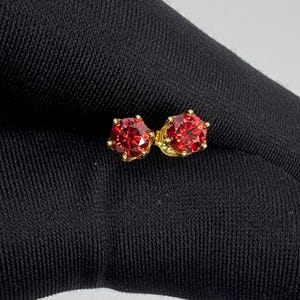 May include: A pair of gold stud earrings, each featuring a large, faceted, round-cut red gemstone. The earrings are set on a black background, highlighting the vibrant color and sparkle of the stones.