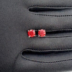 May include: A pair of red gemstone stud earrings with a silver setting. The round-cut gemstones are a vibrant red color. The earrings are displayed on a black surface.