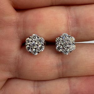 May include: A pair of silver-toned stud earrings, each featuring a cluster of seven round, clear, faceted stones arranged in a floral pattern. The stones are set in a metallic base, creating a bright, reflective appearance on each earring.