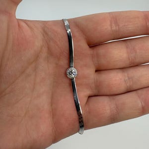 May include: A silver bracelet with a herringbone chain design. The bracelet features a round, clear gemstone in the center, set in a halo of smaller stones. The bracelet is displayed on a person's hand.