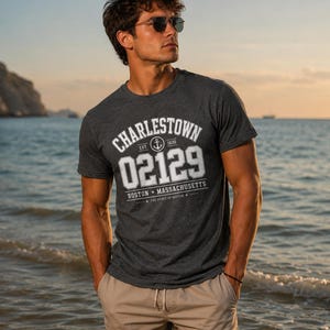 May include: A dark charcoal grey short-sleeved t-shirt featuring white screen-printed text that reads CHARLESTOWN 02129 BOSTON MASSACHUSETTS with an anchor icon and the text EST 1630 and THE SPIRIT OF BOSTON, paired with beige drawstring shorts.