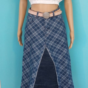 May include: A mannequin wearing a white lace crop top, a denim plaid skirt with a front slit, and a pink belt with a silver buckle. A black hat with red and blue floral and peacock accents completes the outfit.