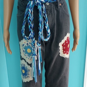 May include: Dark gray denim jeans with decorative floral patches in blue, yellow, and red. A braided blue and white belt is tied around the waist. The jeans have a frayed hem.