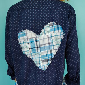 May include: A navy blue long-sleeved shirt with a white and blue plaid heart applique on the back. The shirt has a white and blue plaid ruffle at the bottom. The shirt is made of a soft, comfortable material.