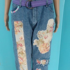 May include: Blue denim jeans with floral fabric patches and a purple belt. The jeans feature decorative floral patches on the front and sides, with a purple belt and a silver buckle. The jeans have a relaxed fit and a frayed hem.