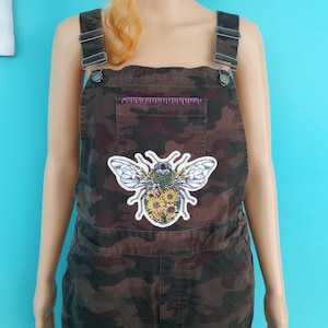May include: Brown camouflage overalls with adjustable straps and a front pocket. A large patch of a bee with floral details is centered on the chest. The overalls are displayed on a mannequin against a turquoise background.