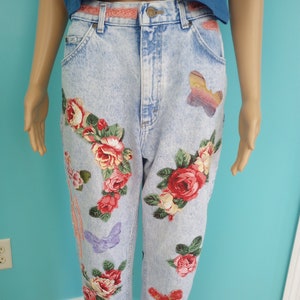 May include: Light wash denim jeans with floral and butterfly appliqués. The jeans feature a high waist, a frayed hem, and a decorative lace belt. The floral designs include red, pink, and white roses with green leaves.