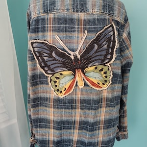 May include: Blue plaid button-up shirt with a large butterfly patch on the back. The butterfly has black, blue, yellow, and red details. The shirt has a classic collar and long sleeves.