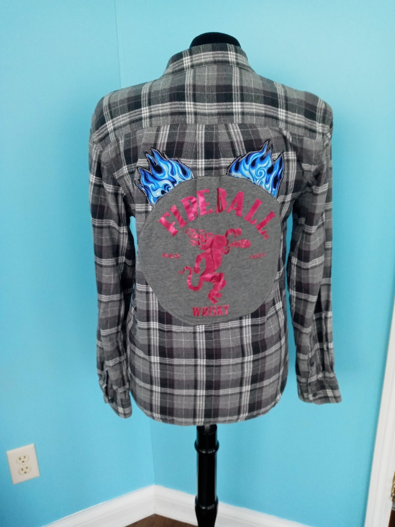 May include: A gray and black plaid flannel shirt with a gray patch on the back. The patch has a pink embroidered design of a monkey with flames on either side. The text "Fireball Whiskey" is written around the monkey.