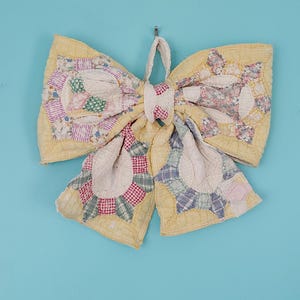 May include: A large decorative bow constructed from quilted fabric featuring a pale yellow base color. The bow is adorned with various patchwork patterns in shades of pink, green, blue, and white, showcasing intricate stitching and fabric textures.