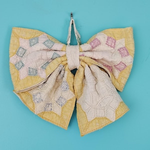 May include: A decorative bow crafted from quilted fabric featuring a yellow border and a central panel of white fabric with stitched star patterns in muted shades of pink, blue, and grey. The bow includes a small fabric loop at the top for hanging.