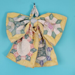 May include: A decorative bow constructed from quilted fabric featuring a patchwork star pattern in shades of yellow, green, white, and purple. The textile surface displays intricate stitching throughout, with a central knot and two hanging tails.