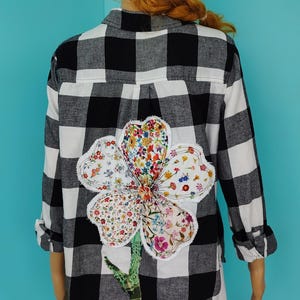 May include: Black and white plaid button-down shirt with a large, colorful floral applique on the back. The flower design is made of various patterned fabrics. The sleeves are rolled up.
