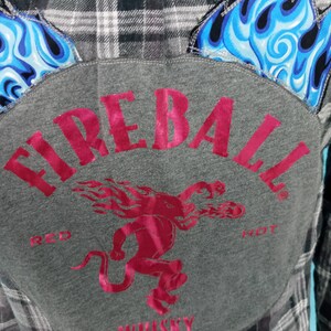 May include: A gray flannel shirt with a patch on the back that says "Fireball Red Hot Whiskey". The patch features a red devil with flames and the words "Red Hot" in white. The shirt has a black and white plaid pattern.