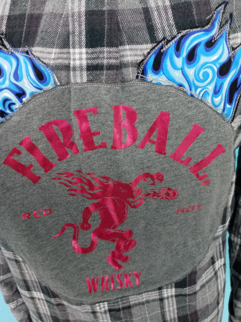 May include: A gray flannel shirt with a patch on the back that says "Fireball Red Hot Whiskey". The patch features a red devil with flames and the words "Red Hot" in white. The shirt has a black and white plaid pattern.