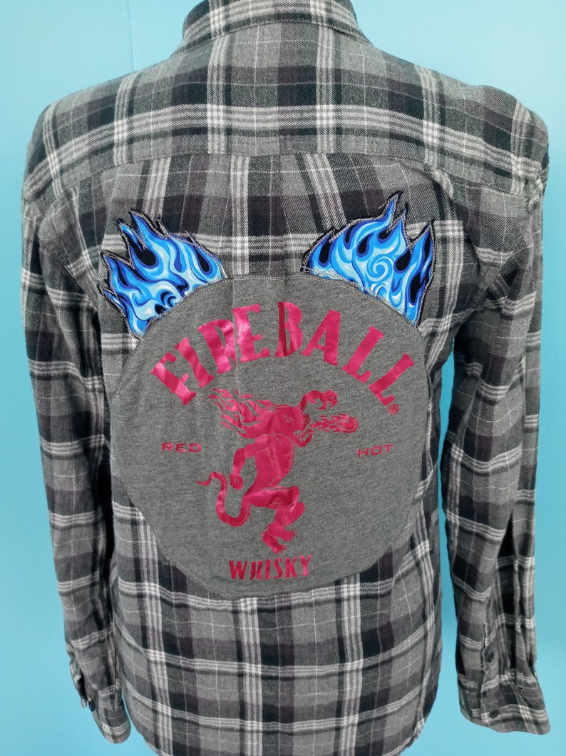 May include: Gray plaid flannel shirt with a gray patch on the back. The patch has a red devil with flames and the text "Fireball Whiskey" in red.