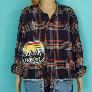 May include: A navy blue plaid shirt with a Wrangler patch featuring a mountain scene. The shirt has two denim pockets and a button-up front. The plaid pattern includes red, yellow, and blue stripes. The shirt is a unique, repurposed garment.