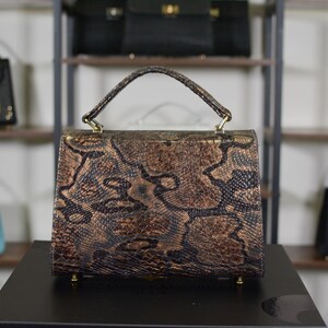 May include: A brown and black snakeskin-patterned handbag with a top handle and gold-coloured hardware. The bag is rectangular with a flap closure. Other handbags are visible on shelves in the background.