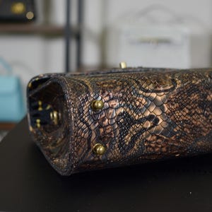 May include: Close-up of a brown and gold snakeskin-patterned handbag. The bag has gold-coloured hardware and a textured surface. The bag is a stylish accessory.