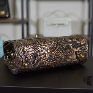 May include: A brown and gold snakeskin-patterned handbag with gold-coloured feet. The bag is rectangular with rounded edges and a gold clasp. Other handbags are visible in the background on shelves.