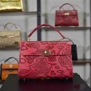 Puede incluir: Un bolso rojo con estampado de piel de serpiente, asa superior y cierre dorado. El bolso se exhibe sobre una superficie negra, con otros bolsos en estantes al fondo. El bolso tiene las iniciales "DM" en el cierre.