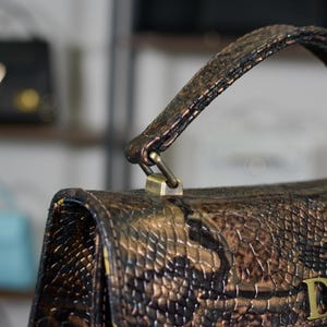 May include: Close-up of a brown and bronze handbag with a snakeskin-effect pattern. The bag features a handle and a gold-coloured clasp. The initials "D&M" are visible on the front.