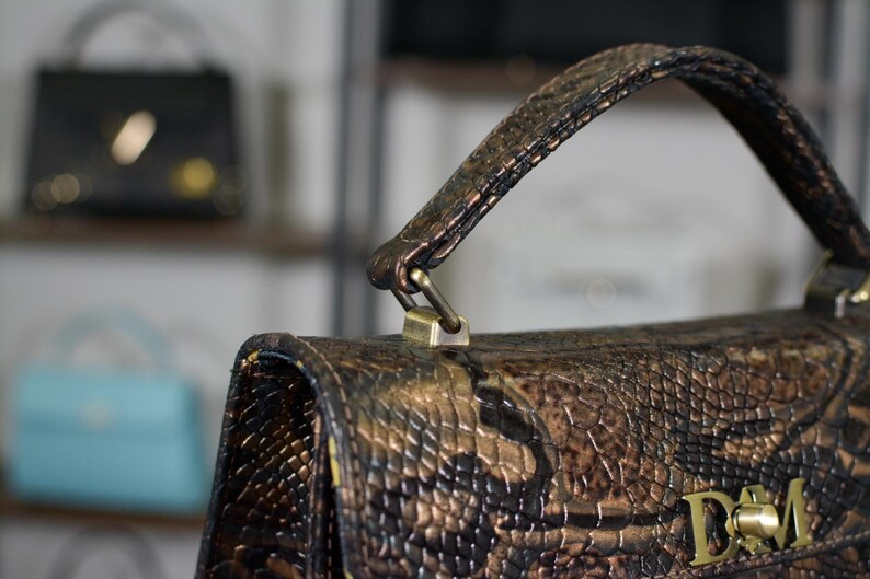 May include: Close-up of a brown and bronze handbag with a snakeskin-effect pattern. The bag features a handle and a gold-coloured clasp. The initials "D&M" are visible on the front.
