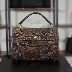 May include: A brown and gold snakeskin-patterned handbag with a top handle and gold hardware. The bag features a flap closure with the initials "DM" and a pearl accent. Other handbags are visible in the background.
