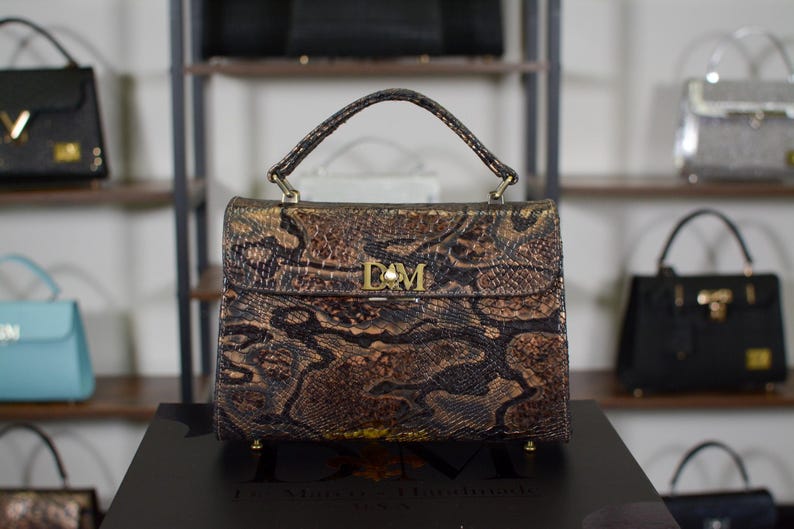 May include: A brown and gold snakeskin-patterned handbag with a top handle and gold hardware. The bag features a flap closure with the initials "DM" and a pearl accent. Other handbags are visible in the background.