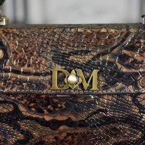 May include: A brown and black snakeskin-patterned handbag with a gold-coloured clasp and handle attachments. The bag features a gold-coloured emblem with the letters "D" and "M". The bag has a textured surface.