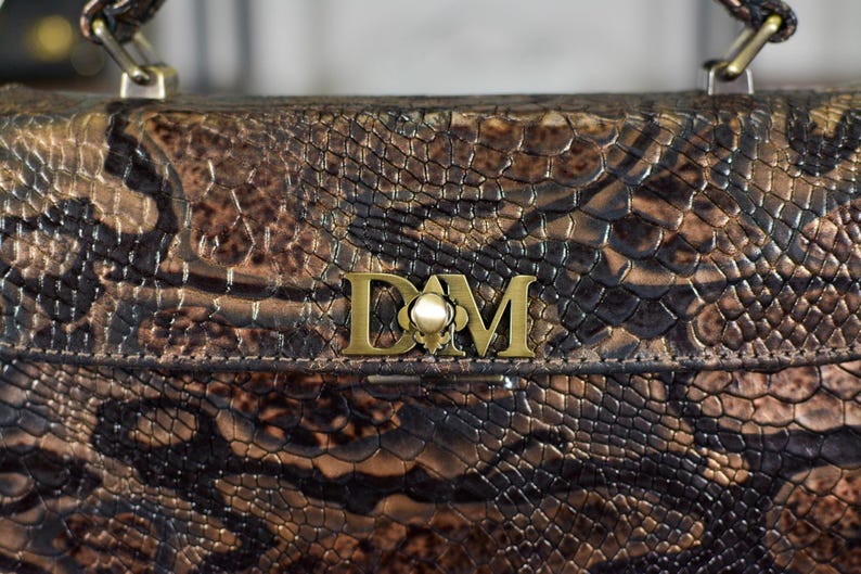May include: A brown and black snakeskin-patterned handbag with a gold-coloured clasp and handle attachments. The bag features a gold-coloured emblem with the letters "D" and "M". The bag has a textured surface.