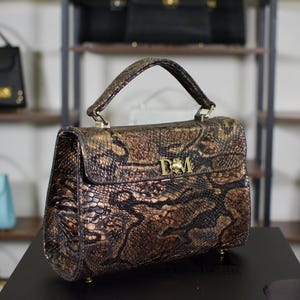 May include: A brown and gold snakeskin-patterned handbag with a top handle and gold hardware. The bag features a flap closure and a gold emblem. Other handbags are visible on shelves in the background.
