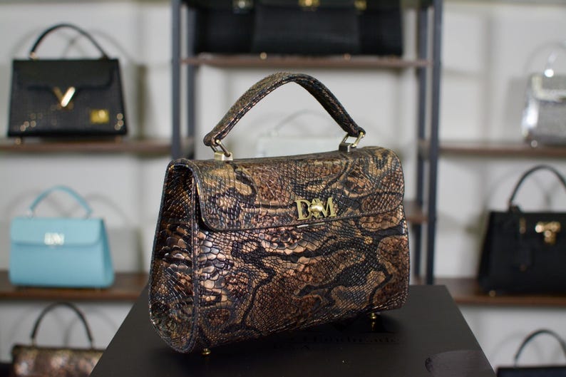 May include: A brown and gold snakeskin-patterned handbag with a top handle and gold hardware. The bag features a flap closure and a gold emblem. Other handbags are visible on shelves in the background.