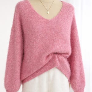 May include: A soft, pink V-neck sweater with long sleeves is displayed on a mannequin. The sweater is paired with white pants. A gold necklace with a small pendant is visible. The sweater has a fluffy texture.