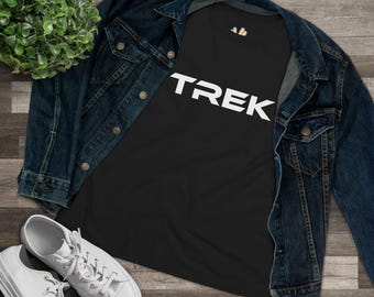 May include: A black t-shirt featuring the word TREK in bold white capital letters across the chest, layered under a dark blue denim jacket, accompanied by a pair of white canvas sneakers and a small green potted plant.