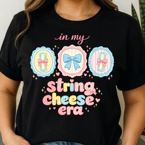 以下が含まれることがあります： 「in my string cheese era」というフレーズがカラフルで遊び心のあるフォントでプリントされた黒いTシャツ。 パステルカラーのリボンとリボンが付いた3つの円形フレームのデザインで、楽しくて気まぐれな外観になっています。